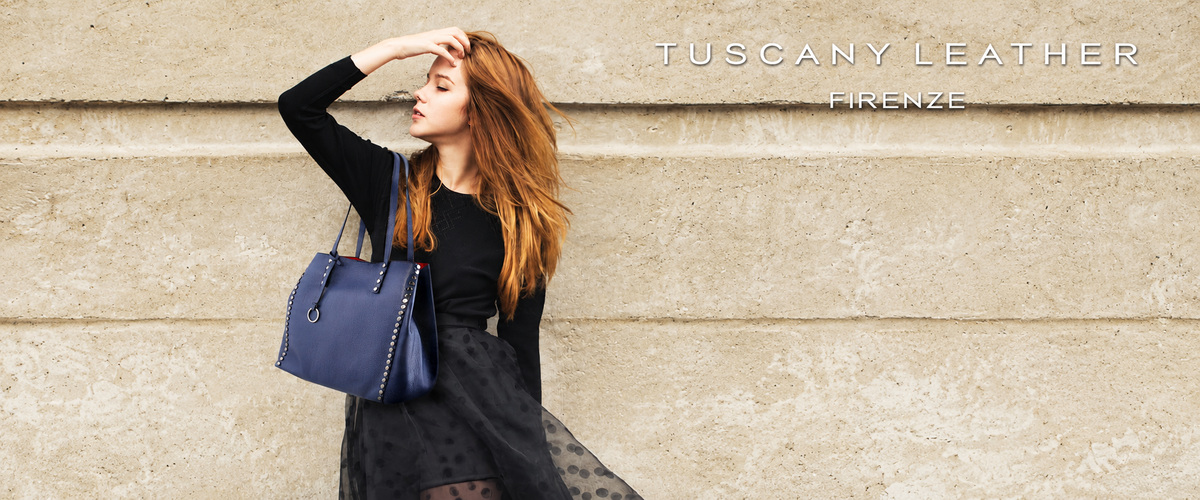 Woman wearing a black outfit and carrying a blue leather tote against a stone wall